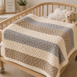May include: A crocheted baby blanket featuring horizontal stripes in alternating shades of cream, light brown, and grey. The blanket has a textured stitch pattern and a solid cream-colored border, draped over the wooden rails of a baby crib.