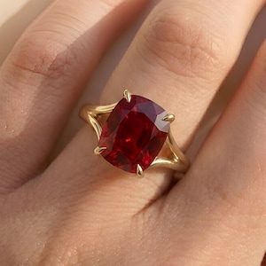 Pode incluir: Um anel de ouro com uma grande pedra preciosa vermelha escura com corte almofada. A banda do anel tem um design dividido, com a pedra preciosa presa por quatro garras. O anel é mostrado no dedo de uma pessoa.