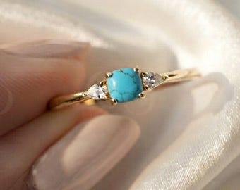 Pode incluir: Um anel de ouro com uma pedra turquesa central de corte almofada, ladeada por duas pequenas pedras claras em forma de pera. A banda é fina e polida, segurando as três pedras numa cravação de garras. O design é simétrico.