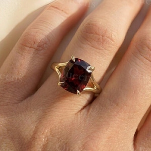 May include: A gold ring featuring a large, cushion-cut deep red garnet gemstone held in a four-prong setting. The band is crafted from polished yellow gold, showcasing a smooth, minimalist design that highlights the dark red stone.