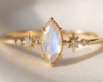 May include: A gold ring featuring a central marquise-cut moonstone set in a beaded bezel. The band is accented on both sides by a small star-shaped setting containing a round clear gemstone, flanked by a tiny round clear gemstone on each outer edge.