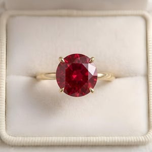 Peut inclure: Une bague en or ornée d'un grand rubis rond et facetté. Le rubis est d'un rouge profond et vibrant, maintenu par quatre griffes en or. La bague présente un design simple et élégant, idéale pour une occasion spéciale.