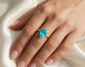 May include: A gold ring featuring a single pear-shaped, bright turquoise-blue gemstone set in a prong setting. The band is thin and polished gold, resting on a finger against a soft, draped white fabric background with a subtle watermark.