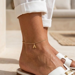 Puede incluir: Tobillera dorada con una cadena delicada y un colgante con la letra "A". La tobillera se lleva en el tobillo, combinada con sandalias blancas. Una joya sencilla y elegante.