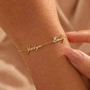 May include: A gold-colored bracelet with the names "Jadyn" and "Emily" in cursive script. The bracelet is delicate, with a chain-link design. The names are connected by the chain. The bracelet is worn on a wrist.
