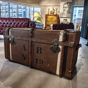 May include: A vintage brown trunk with metal hardware and white stripes. The trunk has the letters "A.N." and "B. 19." printed on the front. The trunk is rectangular and appears to be made of wood and leather.