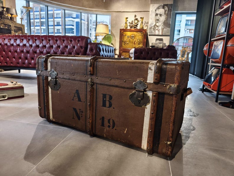 May include: A vintage brown trunk with metal hardware and white stripes. The trunk has the letters "A.N." and "B. 19." printed on the front. The trunk is rectangular and appears to be made of wood and leather.