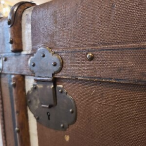 May include: Close-up of a vintage brown trunk with metal hardware. The trunk features a textured canvas exterior, metal clasps, and decorative metal accents. The trunk's design includes vertical white and brown stripes.