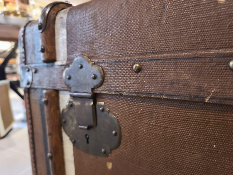 May include: Close-up of a vintage brown trunk with metal hardware. The trunk features a textured canvas exterior, metal clasps, and decorative metal accents. The trunk's design includes vertical white and brown stripes.