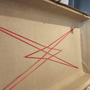 May include: Open vintage trunk interior with tan fabric lining and red ribbon crisscross design. The trunk has a wooden frame and metal hardware. The red ribbon is arranged in a geometric pattern, adding a decorative element to the trunk's interior.