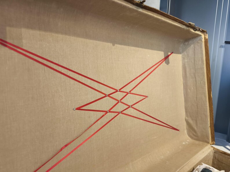 May include: Open vintage trunk interior with tan fabric lining and red ribbon crisscross design. The trunk has a wooden frame and metal hardware. The red ribbon is arranged in a geometric pattern, adding a decorative element to the trunk's interior.