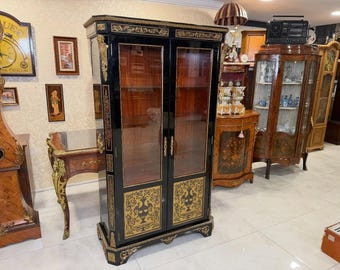 May include: A tall, black antique cabinet with glass doors, gold accents, and decorative panels. The cabinet features ornate carvings and a rich, dark wood interior. Other antique furniture pieces are visible in the background.