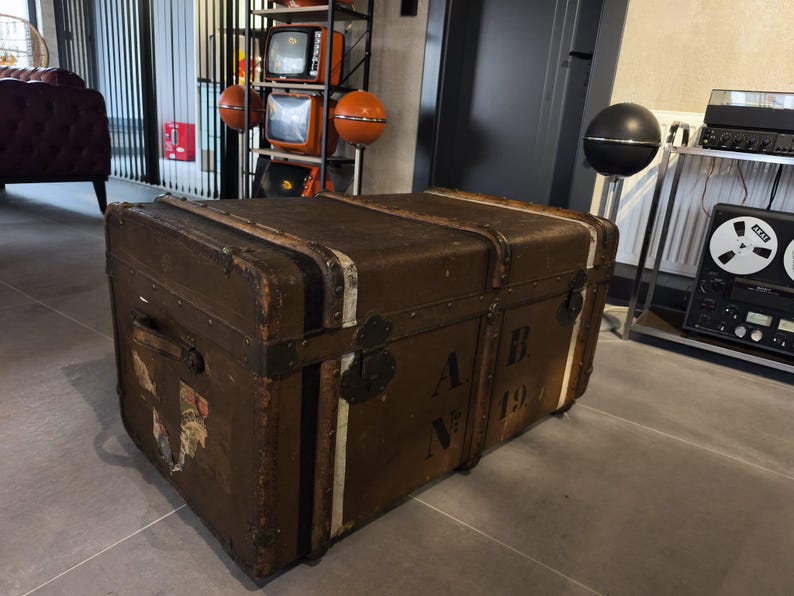 May include: A vintage brown trunk with metal hardware and white stripes. The trunk has the letters "A B" and the number "19" on the side. The trunk is rectangular and appears to be made of wood or a similar material.