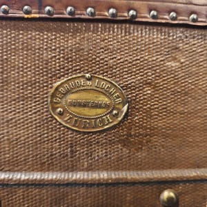 May include: Close-up of a vintage brown leather trunk with a textured surface. The trunk features a brass oval plaque with the text "GEBRUDER LOCHER, KUTZENSTR, ZURICH". The trunk is adorned with metal studs and a leather trim.