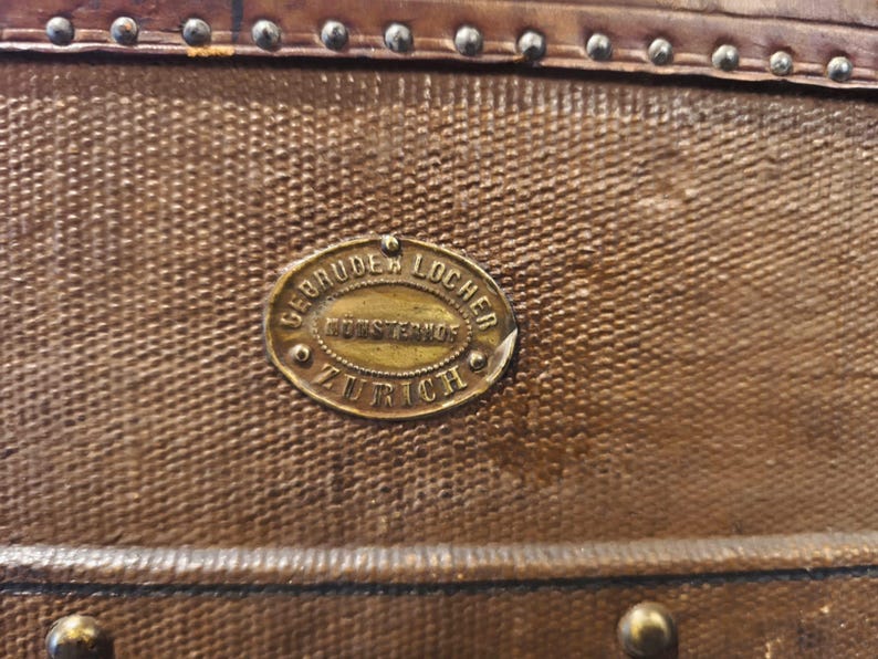 May include: Close-up of a vintage brown leather trunk with a textured surface. The trunk features a brass oval plaque with the text "GEBRUDER LOCHER, KUTZENSTR, ZURICH". The trunk is adorned with metal studs and a leather trim.