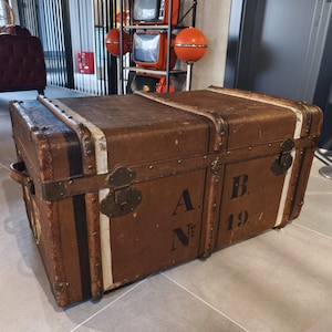 May include: A vintage brown trunk with metal hardware, black and white stripes, and the letters "A B" and the number "194" stenciled on the front. The trunk has a rectangular shape and a handle on the side.
