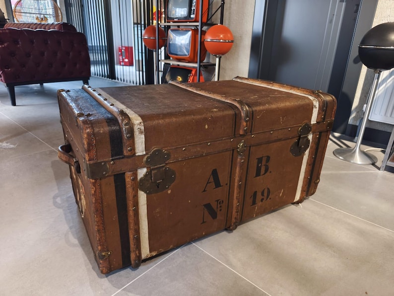 May include: A vintage brown trunk with metal hardware, black and white stripes, and the letters "A B" and the number "194" stenciled on the front. The trunk has a rectangular shape and a handle on the side.