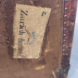 May include: Close-up of a vintage brown travel trunk with a paper label. The label reads "Z&uuml;rich-Hauptbahnhof" with additional text and a blue stamp. The trunk has metal corner protectors and a leather trim with metal studs.