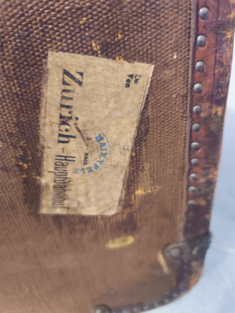 May include: Close-up of a vintage brown travel trunk with a paper label. The label reads "Z&uuml;rich-Hauptbahnhof" with additional text and a blue stamp. The trunk has metal corner protectors and a leather trim with metal studs.
