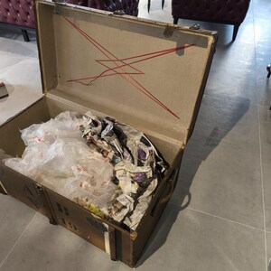May include: An open, vintage brown trunk with a tan interior and red decorative lines. The trunk is filled with crumpled newspaper and clear plastic. The trunk has metal hardware and is sitting on a gray floor.