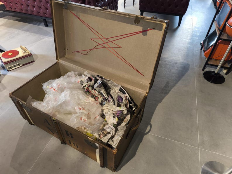 May include: An open, vintage brown trunk with a tan interior and red decorative lines. The trunk is filled with crumpled newspaper and clear plastic. The trunk has metal hardware and is sitting on a gray floor.
