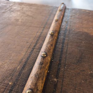 May include: Close-up of a vintage trunk detail. A weathered wooden strip with brass-colored studs runs diagonally across the textured, brown fabric of the trunk's surface. The wood shows signs of age and wear, adding to the antique aesthetic.