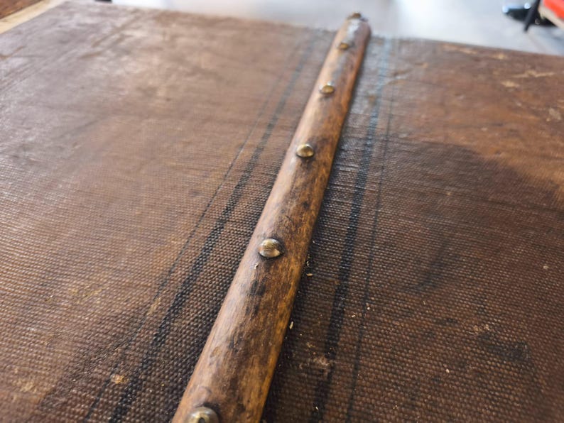 May include: Close-up of a vintage trunk detail. A weathered wooden strip with brass-colored studs runs diagonally across the textured, brown fabric of the trunk's surface. The wood shows signs of age and wear, adding to the antique aesthetic.