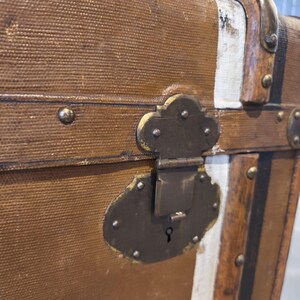 May include: Close-up of a vintage brown trunk with metal hardware. The trunk features a textured canvas exterior, metal corner protectors, and a decorative lock. The trunk has white and black accents and is likely used for travel or storage.