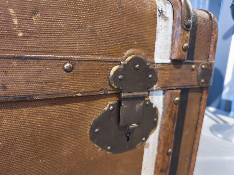 May include: Close-up of a vintage brown trunk with metal hardware. The trunk features a textured canvas exterior, metal corner protectors, and a decorative lock. The trunk has white and black accents and is likely used for travel or storage.