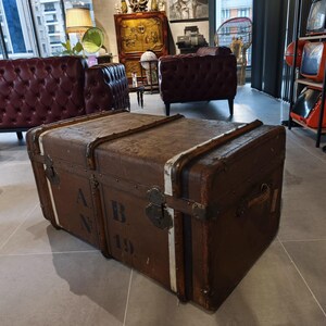 May include: A large, vintage brown trunk with metal hardware and white trim. The trunk has the letters "A.B. 19" stenciled on the side. The trunk is a piece of antique furniture.