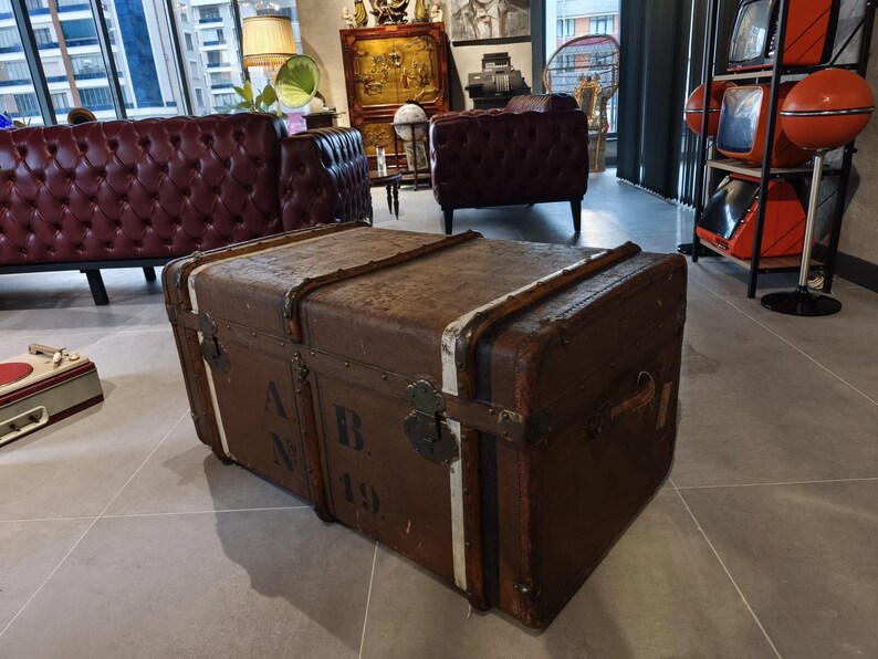 May include: A large, vintage brown trunk with metal hardware and white trim. The trunk has the letters "A.B. 19" stenciled on the side. The trunk is a piece of antique furniture.