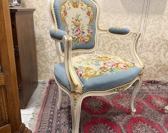May include: An antique armchair with a light blue upholstered seat and back, adorned with floral embroidery. The chair has a white wooden frame with gold accents and curved armrests. The chair is a vintage piece.