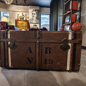 May include: Two antique brown trunks with metal hardware and white stripes. The trunks have the letters "A.N." and "B. 19." printed on them. The trunks are likely used for storage or decoration.