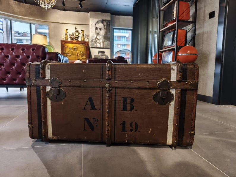 May include: Two antique brown trunks with metal hardware and white stripes. The trunks have the letters "A.N." and "B. 19." printed on them. The trunks are likely used for storage or decoration.