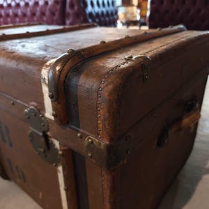 May include: A vintage brown trunk with metal hardware and corner protectors. The trunk has a rectangular shape with a slightly curved top. The trunk has a leather handle and metal latches. The trunk is made of wood and has a distressed finish.