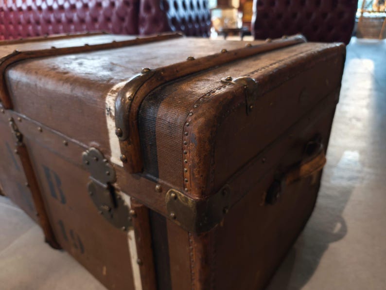 May include: A vintage brown trunk with metal hardware and corner protectors. The trunk has a rectangular shape with a slightly curved top. The trunk has a leather handle and metal latches. The trunk is made of wood and has a distressed finish.