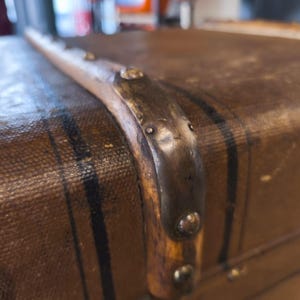 May include: Close-up of a vintage brown trunk with black stripes and metal corner reinforcements. The trunk's surface has a textured appearance, and the metal accents show signs of age.