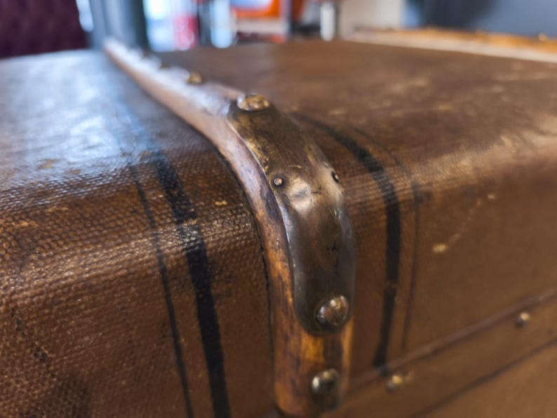 May include: Close-up of a vintage brown trunk with black stripes and metal corner reinforcements. The trunk's surface has a textured appearance, and the metal accents show signs of age.