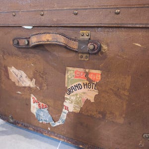 May include: Vintage brown travel trunk with leather handle and metal hardware. The trunk features several aged travel stickers, including one that reads "Grand Hotel". The trunk is rectangular and has metal corner protectors.
