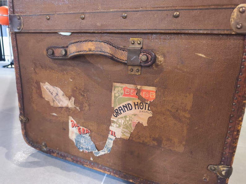 May include: Vintage brown travel trunk with leather handle and metal hardware. The trunk features several aged travel stickers, including one that reads "Grand Hotel". The trunk is rectangular and has metal corner protectors.