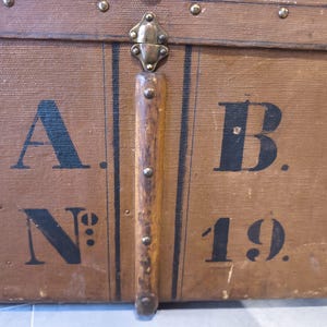 May include: Close-up of a vintage brown trunk with black lettering and metal hardware. The trunk features the letters "A. B. N&deg; 19." and is accented with dark vertical stripes and metal studs.
