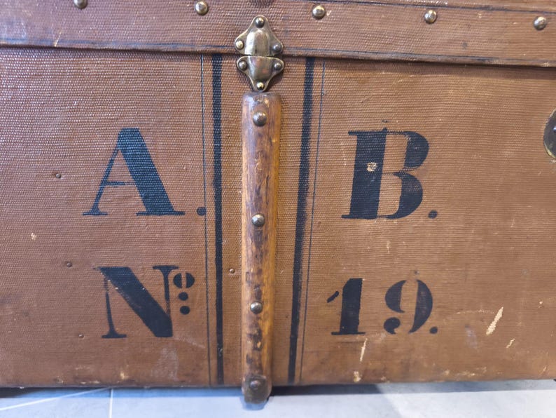 May include: Close-up of a vintage brown trunk with black lettering and metal hardware. The trunk features the letters "A. B. N&deg; 19." and is accented with dark vertical stripes and metal studs.