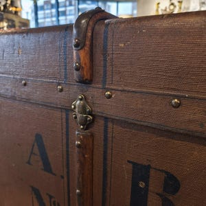 May include: Close-up of a vintage brown trunk with metal hardware and leather accents. The trunk features a lock, corner protectors, and decorative rivets. The trunk has the letter "A" and "B" printed on the side.