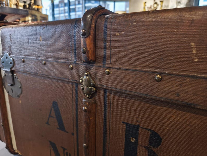 May include: Close-up of a vintage brown trunk with metal hardware and leather accents. The trunk features a lock, corner protectors, and decorative rivets. The trunk has the letter "A" and "B" printed on the side.