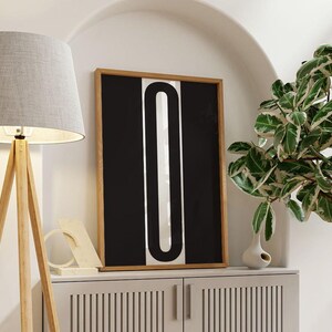 May include: A framed abstract art print featuring a black background with a white, vertical, U-shaped design. The artwork is displayed on a white cabinet, next to a wooden floor lamp with a gray lampshade and a potted plant.