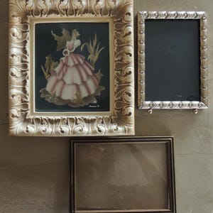 May include: A collection of three picture frames: one square cream-colored frame with ornate scrollwork containing a print of a figure in a ruffled dress, one rectangular silver-toned frame with a textured border, and one simple dark brown rectangular frame.
