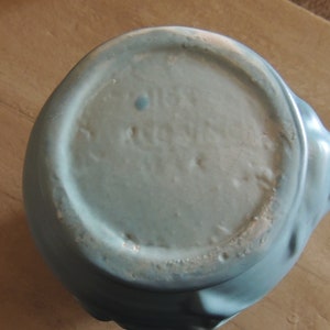 May include: The bottom of a light blue ceramic teapot with a smooth, glazed finish. The teapot has a rounded body and a spout on the side.