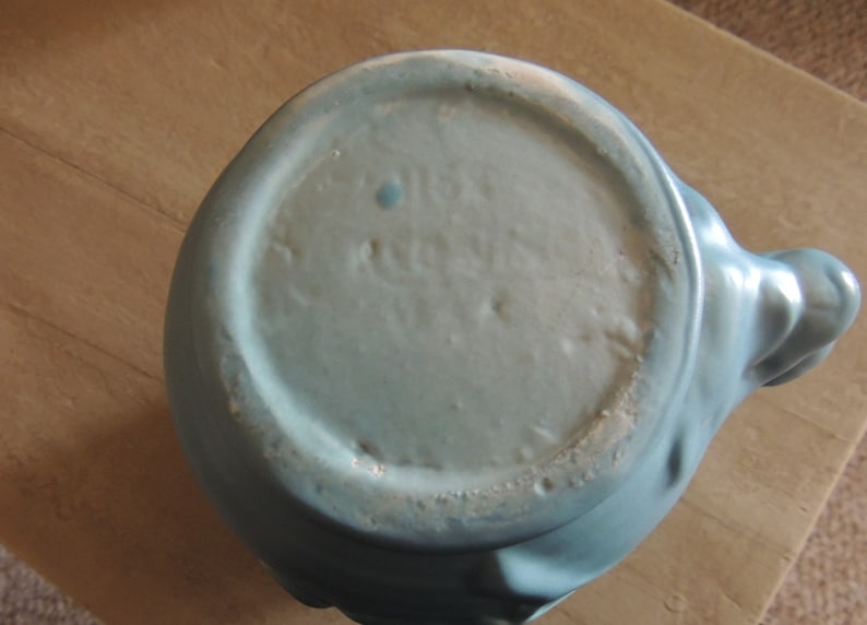 May include: The bottom of a light blue ceramic teapot with a smooth, glazed finish. The teapot has a rounded body and a spout on the side.