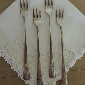 May include: Four silver forks with ornate floral detailing on the handles are arranged on a white, lace-edged napkin. The forks have three tines each and are set against a light brown wooden surface.