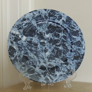 May include: A blue and white marble charger plate with a raised rim. The plate is on a clear plastic stand.